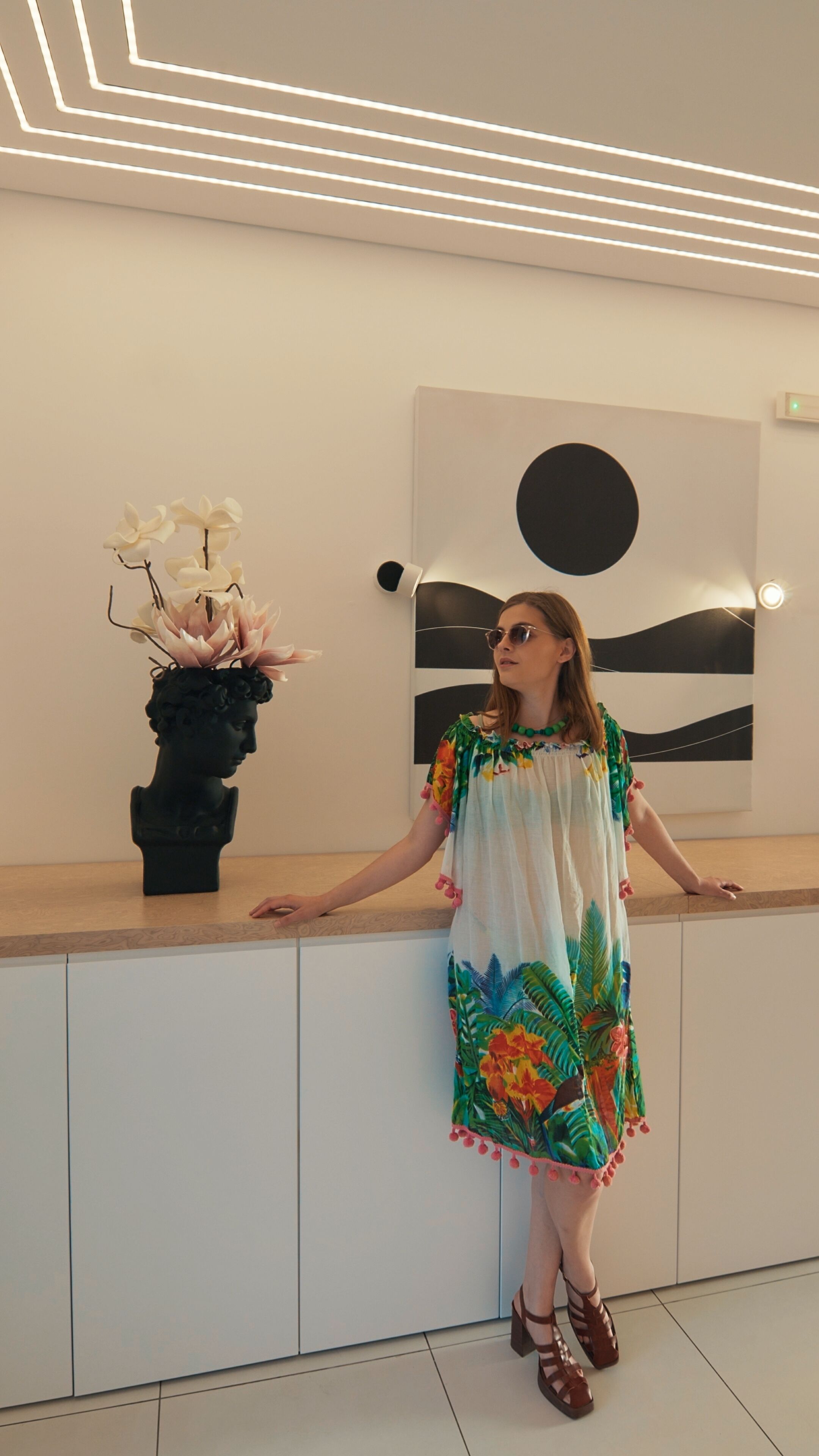 A person standing in a modern room with a minimalist design, featuring a wooden counter, a black bust sculpture, and a wall-mounted artwork.