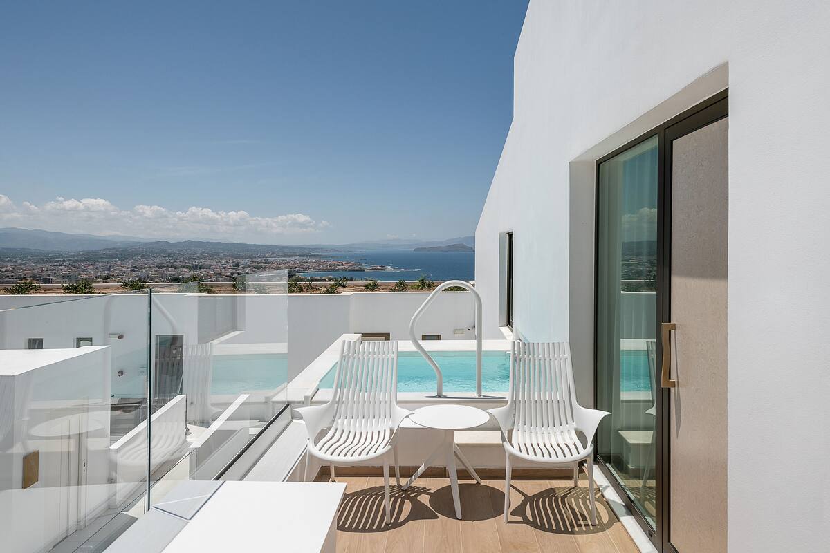 Balcón con piscina, dos sillas blancas y baranda de cristal con vistas a la ciudad y al mar.