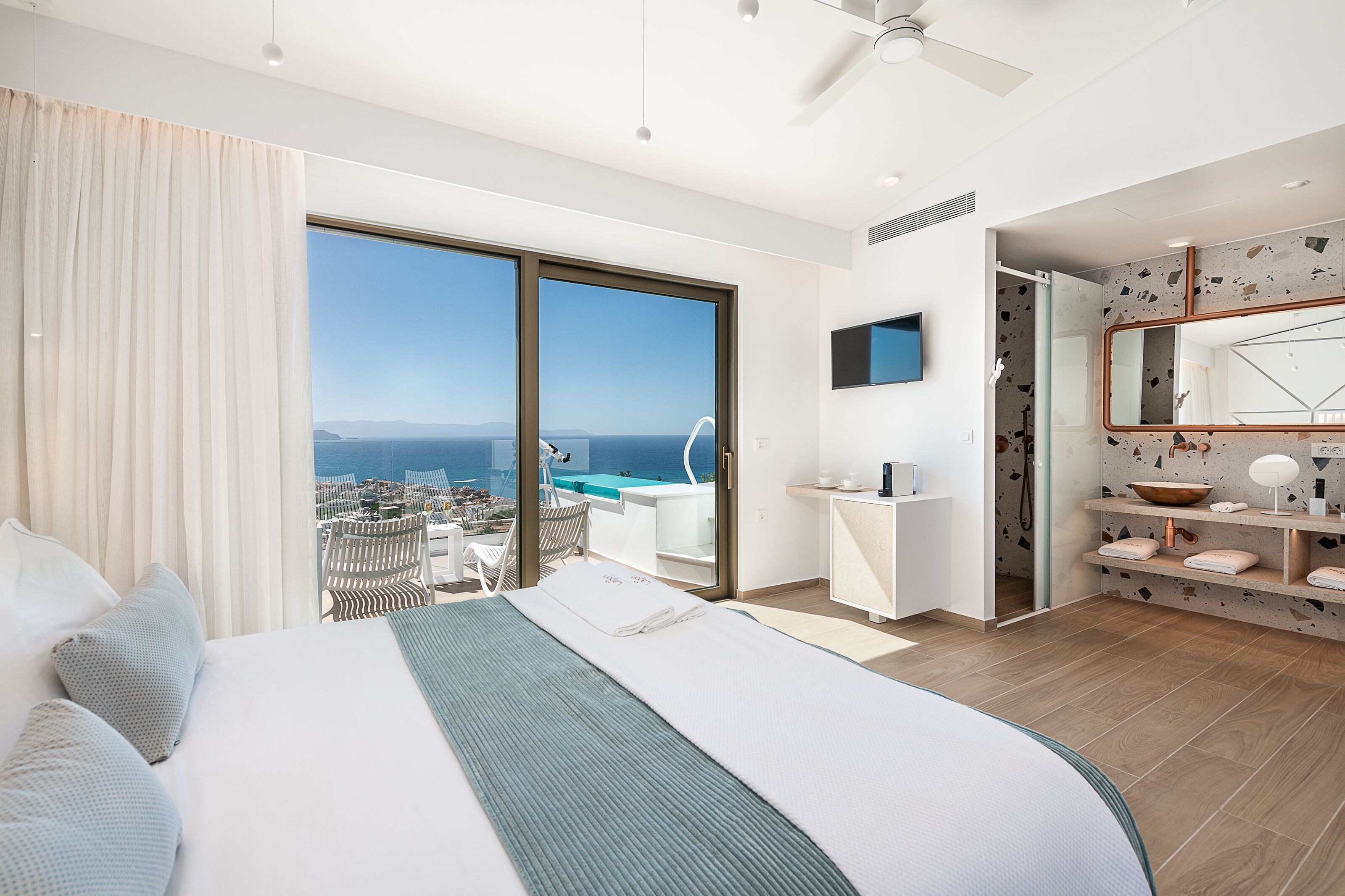 A modern hotel room with a large bed, a bathroom with a shower, and a balcony with a view of the sea.