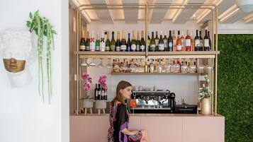 Zona de bar con una mujer sentada en un taburete, rodeada de sillas blancas, y una estantería con vinos y cócteles detrás.