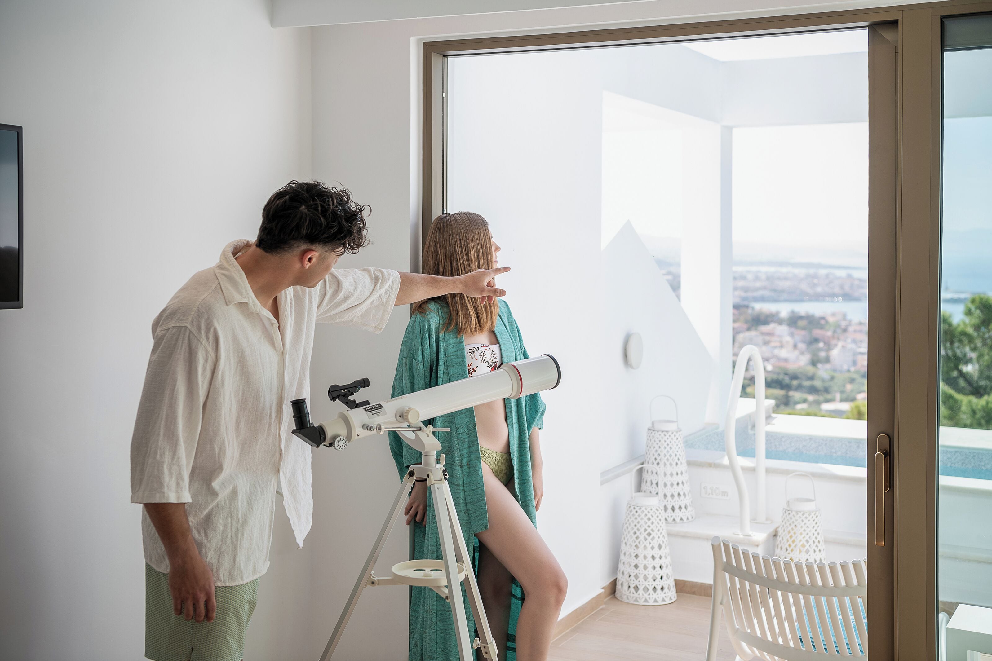 Ein Zimmer mit einem Teleskop, ein Balkon mit Blick auf eine Stadtlandschaft und ein weißer Stuhl.
