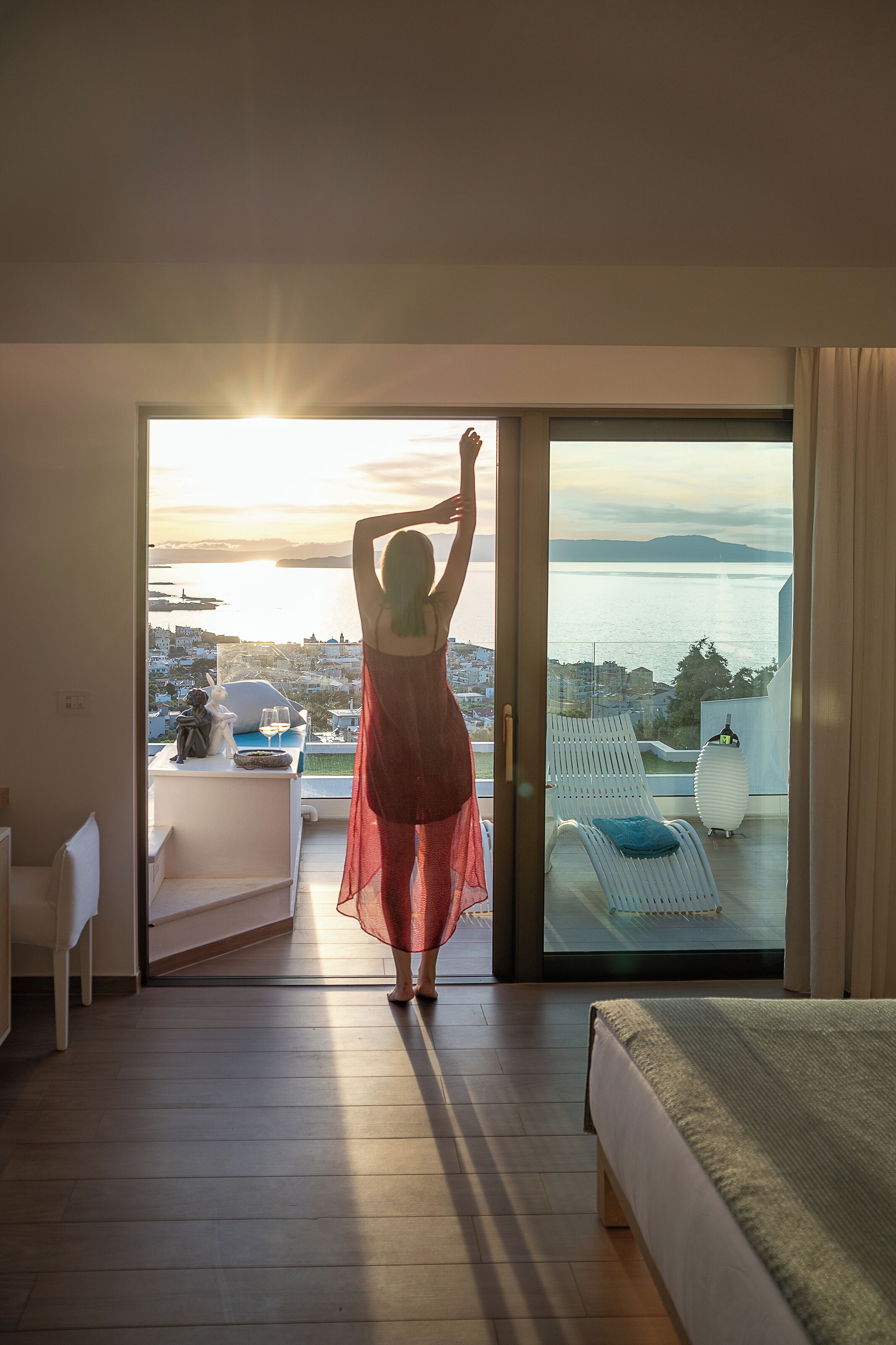 A woman in a red dress standing in a doorway with a view of a balcony and a cityscape.