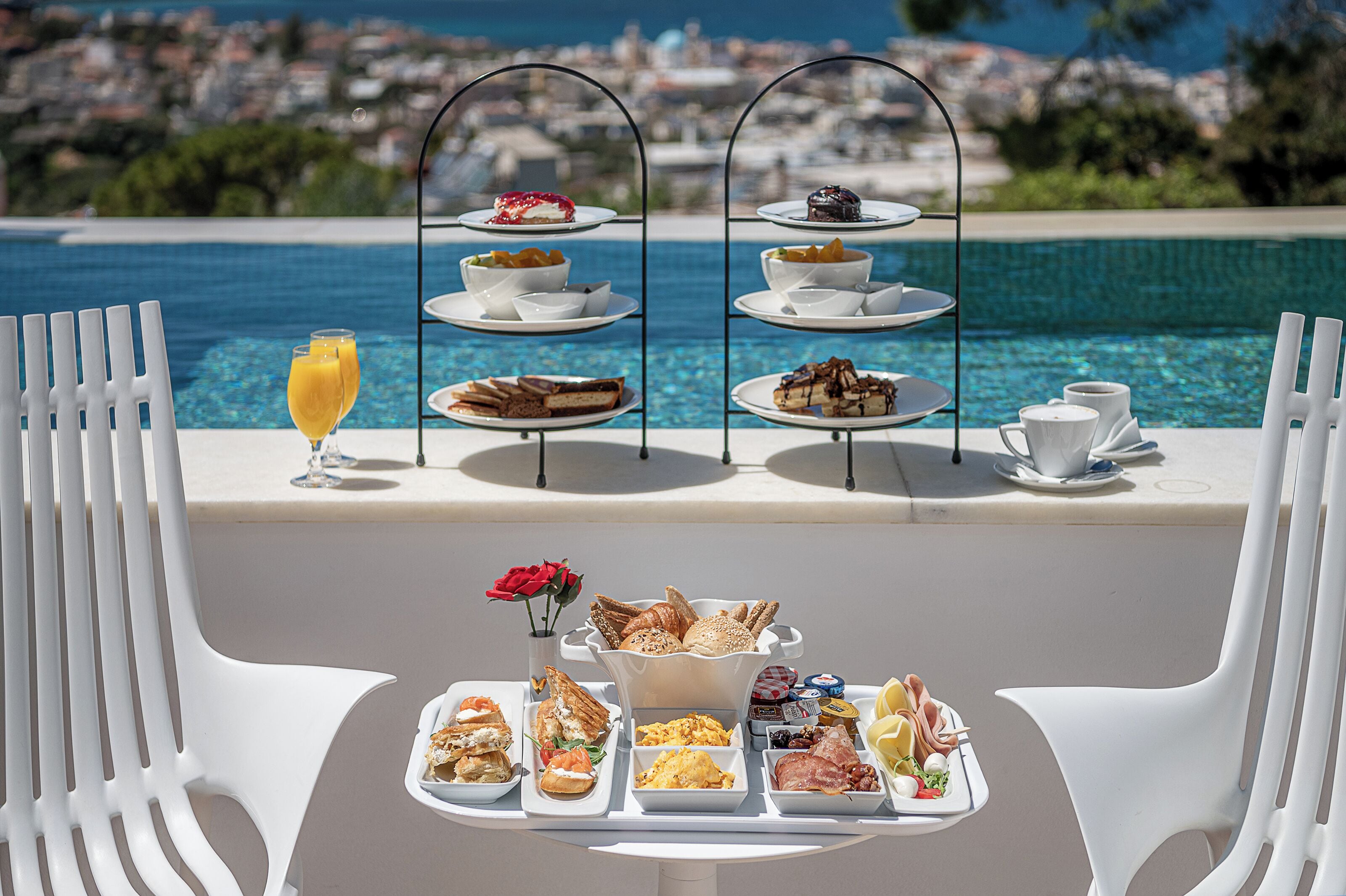 Un espace de restauration extérieur baigné de soleil, avec vue sur la mer, comprenant un présentoir à trois niveaux pour les desserts, un plateau de produits pour le petit-déjeuner et des chaises en plastique blanches.