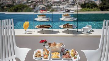 Un espace de restauration extérieur baigné de soleil, avec vue sur la mer, comprenant un présentoir à trois niveaux pour les desserts, un plateau de produits pour le petit-déjeuner et des chaises en plastique blanches.