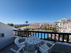 A balcony with a table and chairs, overlooking a pool and a resort complex with multiple buildings and palm trees. - Golden View Holiday Home (Playa de las Américas)