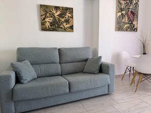 Een grijze bank met kussens, een witte tafel met stoelen, en twee kunstwerken aan de muur.