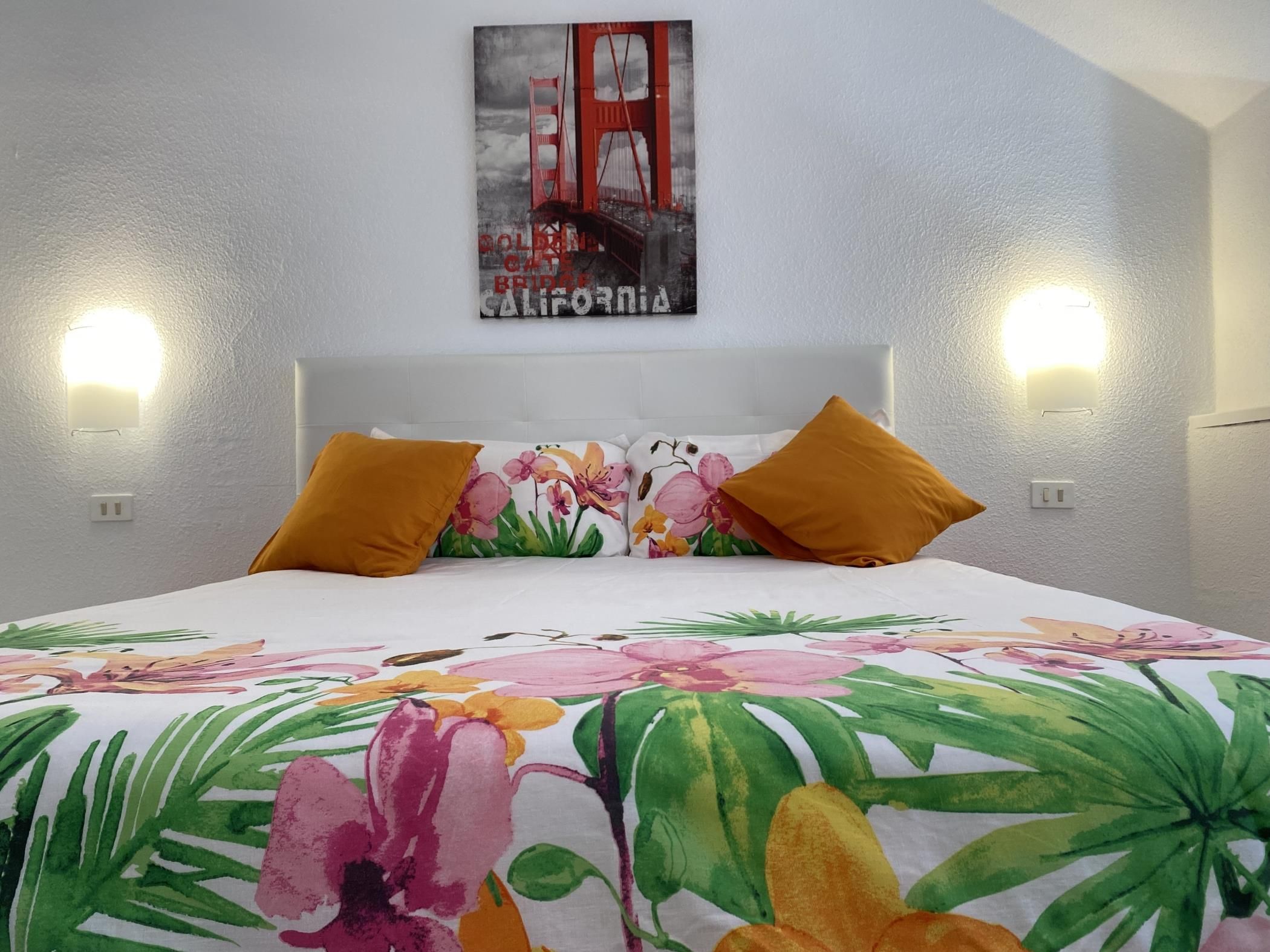 A hotel room with a bed, floral bedspread, two wall-mounted lights, and a painting of the Golden Gate Bridge.
