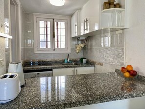 Een keuken met granieten aanrechtbladen, witte kasten, een inbouwoven en een fruitschaal op het aanrecht.