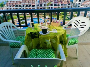 A balcony with a table set for two, chairs with green polka dot cushions, and a view of a resort with buildings and a pool.