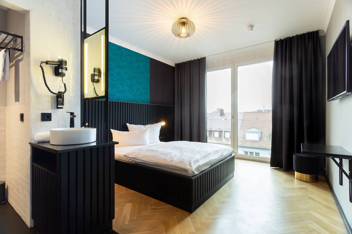 A modern hotel room with a large bed, a bedside table, a built-in sink, a mirror, and a large window with curtains.