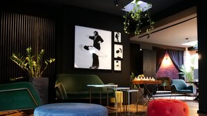 A modern lounge area with a green sofa, a blue ottoman, and a red pouf. There are framed pictures on the wall and a plant in the corner.