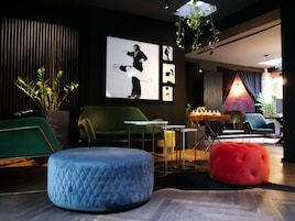 A modern lounge area with a green sofa, a blue ottoman, and a red pouf. There are framed pictures on the wall and a plant in the corner.