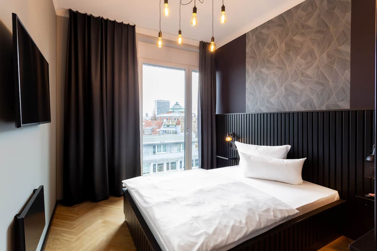 A modern hotel room with a large bed, a flat-screen TV, and a city view through the window.