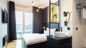 A modern hotel room with a large bed, a vanity with a mirror, a hairdryer, and a view of the city through the window.