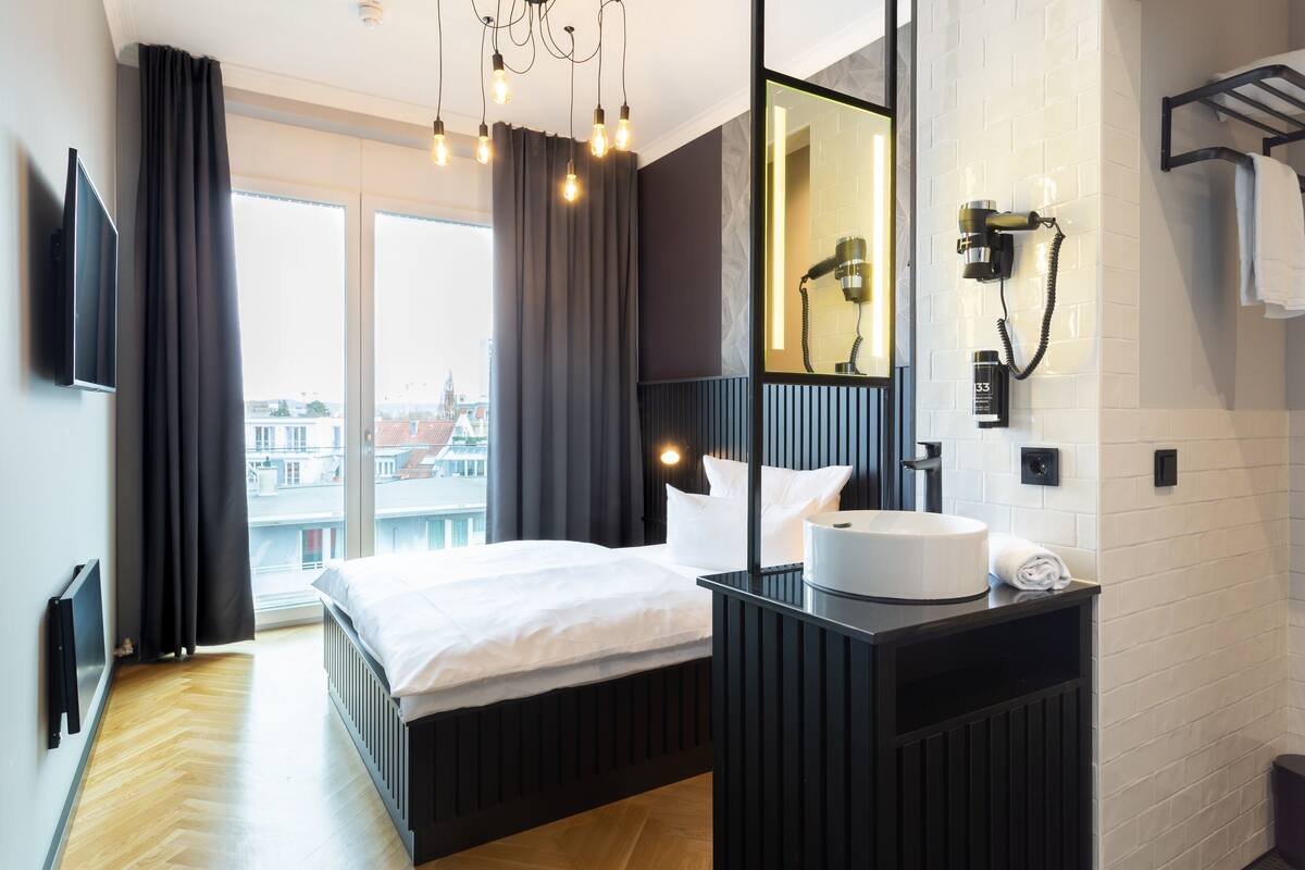 A modern hotel room with a large bed, a vanity with a mirror, a hairdryer, and a view of the city through the window.
