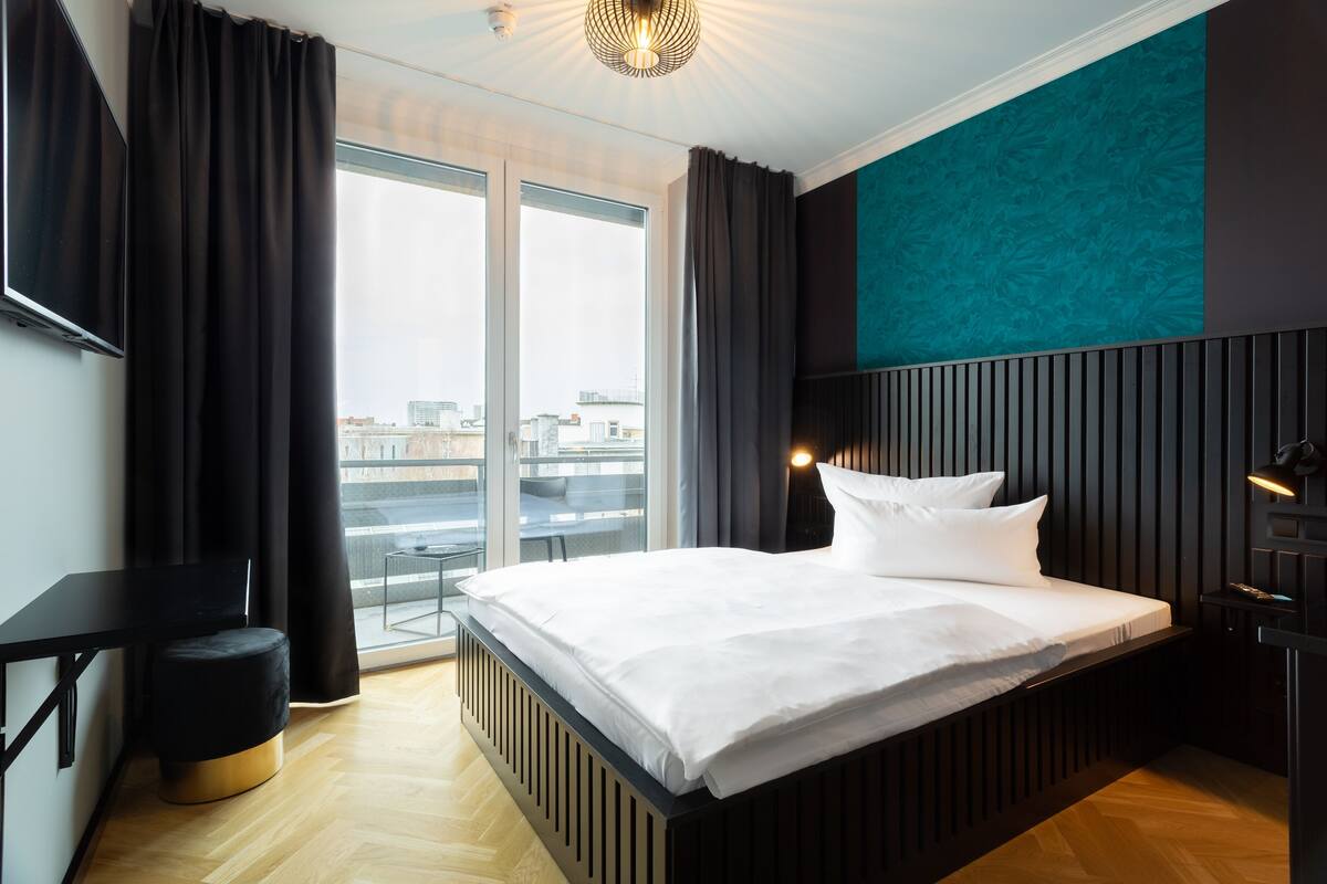 A modern hotel room with a large bed, a desk, and a view of the city through large windows.