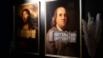 Two framed artworks on a wall, one depicting a classical painting with a figure and the other a portrait of a historical figure with currency notes superimposed.