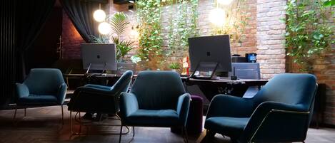 Un espacio de trabajo moderno con dos escritorios, dos sillas azules y plantas colgantes frente a una pared de ladrillo.