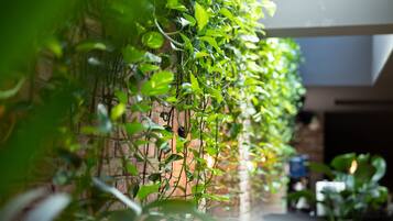 A vertical garden with lush green plants against a brick wall, a glass table in the foreground, and a glimpse of a room with a bed and a nightstand.