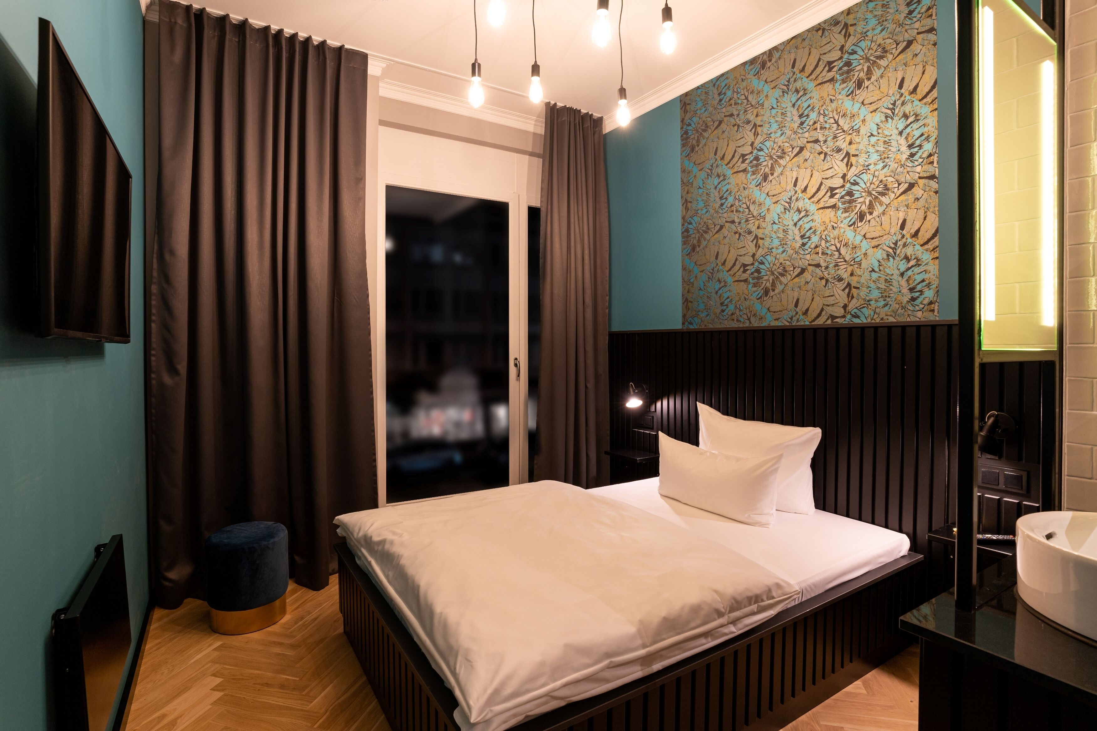 A modern bedroom with a large bed, a small stool, a wall-mounted TV, and a bathroom with a sink and mirror.