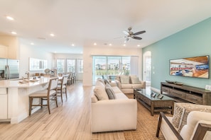 A spacious living area with a large sofa, a coffee table, and a TV on the wall. There is a kitchen area with a fridge and a dining table.