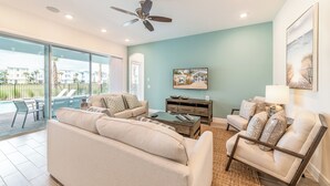 A spacious living room with a large sofa, a coffee table, and a chair. There is a ceiling fan, a painting on the wall, and a view of the outside through large windows.
