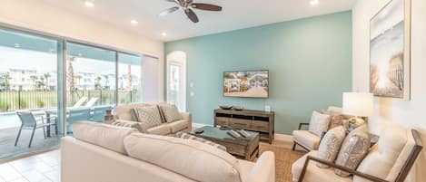 A spacious living room with a large sofa, a coffee table, and a chair. There is a ceiling fan, a painting on the wall, and a view of the outside through large windows.