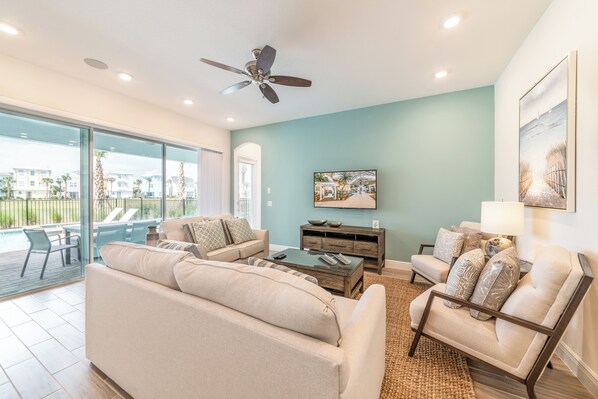 A spacious living room with a large sofa, a coffee table, and a chair. There is a ceiling fan, a painting on the wall, and a view of the outside through large windows.