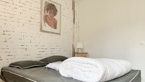 A bed with a grey and white duvet, two pillows, a nightstand with a lamp, and a painting on a white brick wall.