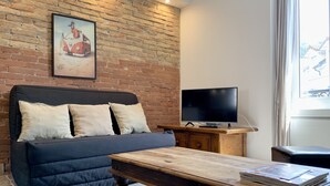 A living room with a brick wall, a sofa with cushions, a television on a wooden stand, and a coffee table with magazines.