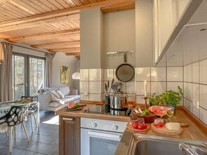 Une cuisine moderne avec un plafond en bois, un coin repas avec une table et des chaises blanches, et une vue sur l’extérieur grâce à de grandes fenêtres.