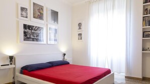 Une chambre avec un lit blanc, une literie rouge et une tête de lit blanche. On peut voir des tableaux encadrés au mur, une table de chevet avec une lampe et une bibliothèque.
