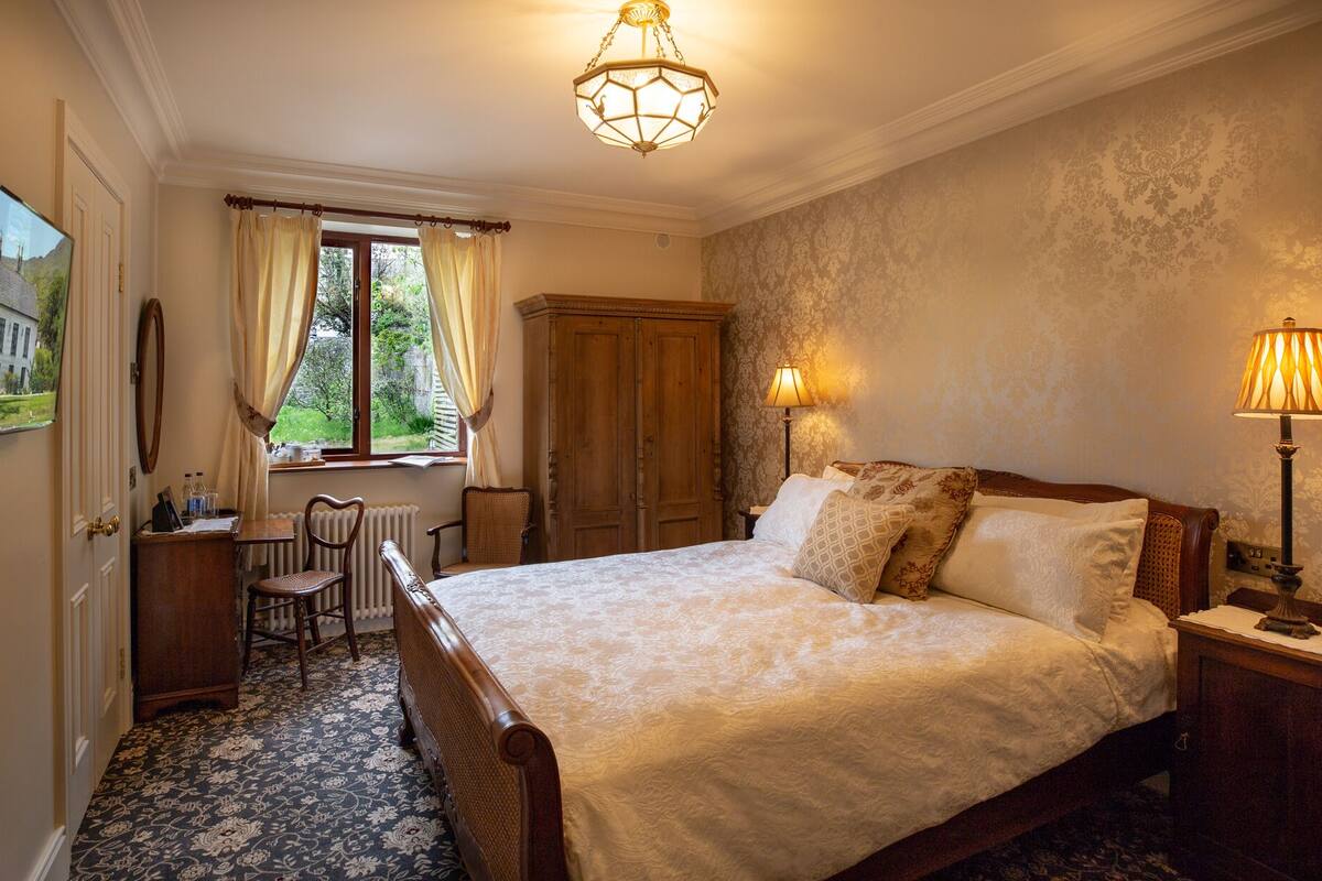 A bedroom with a large bed, a wooden wardrobe, a desk with a chair, a radiator, and a window with curtains.