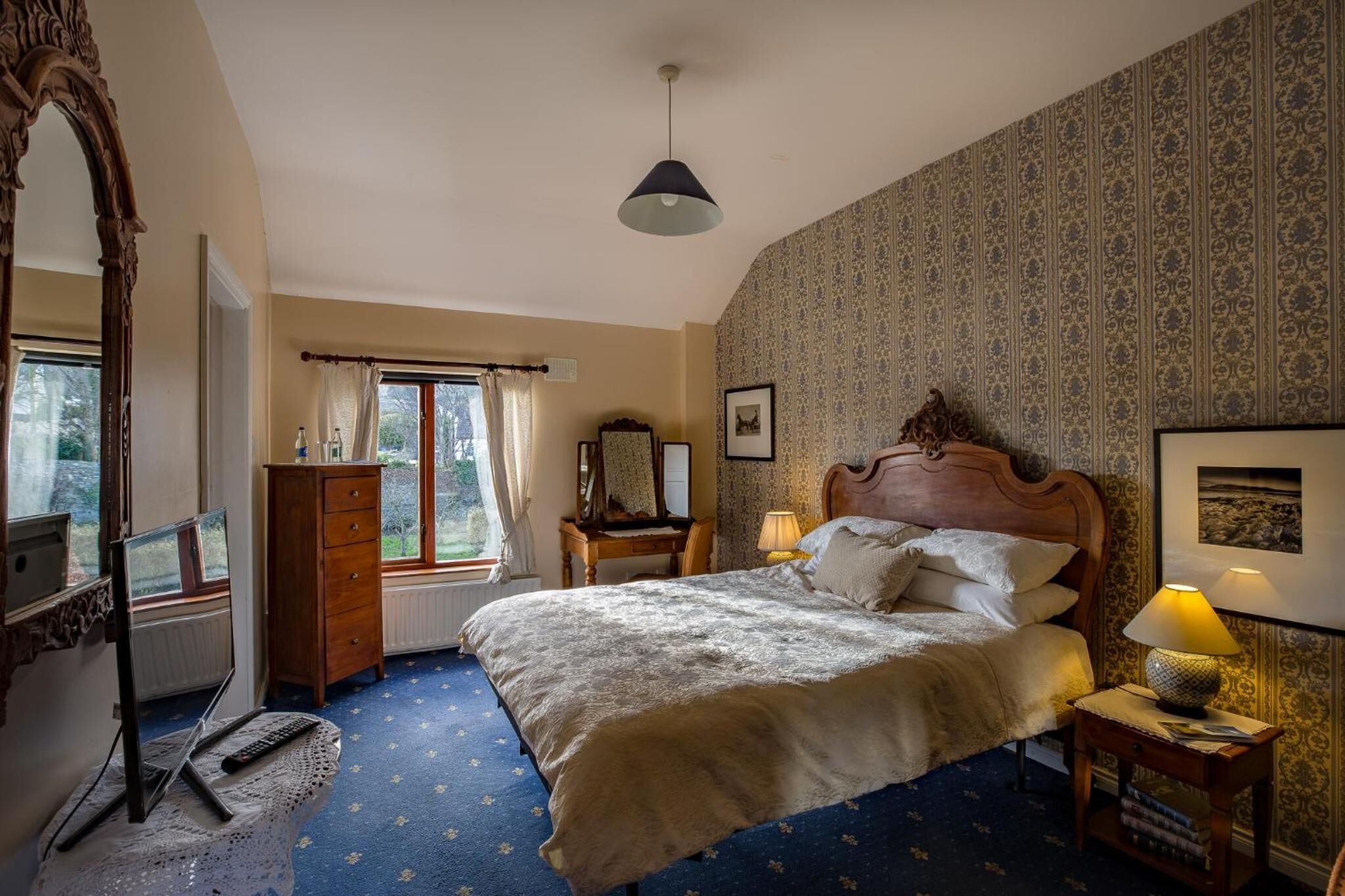A bedroom with a large bed, a wooden dresser, a desk with a lamp, a mirror, and a window with curtains.