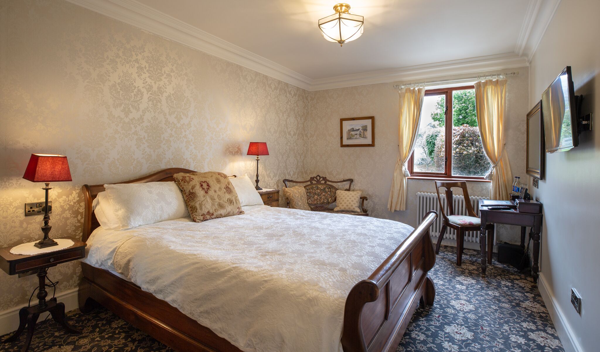 Quarto com cama grande, duas mesas de cabeceira com luminárias, uma cadeira de madeira, escrivaninha e janela com cortinas.