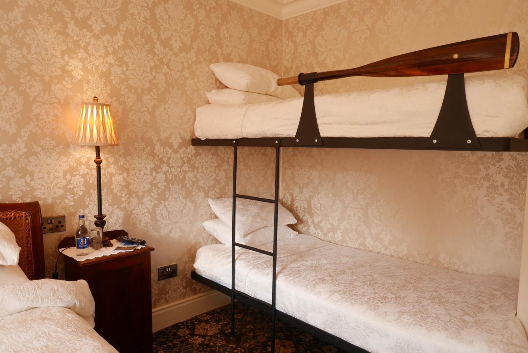 A hotel room with a bunk bed, a bedside table with a lamp, a bottle, and a remote control.
