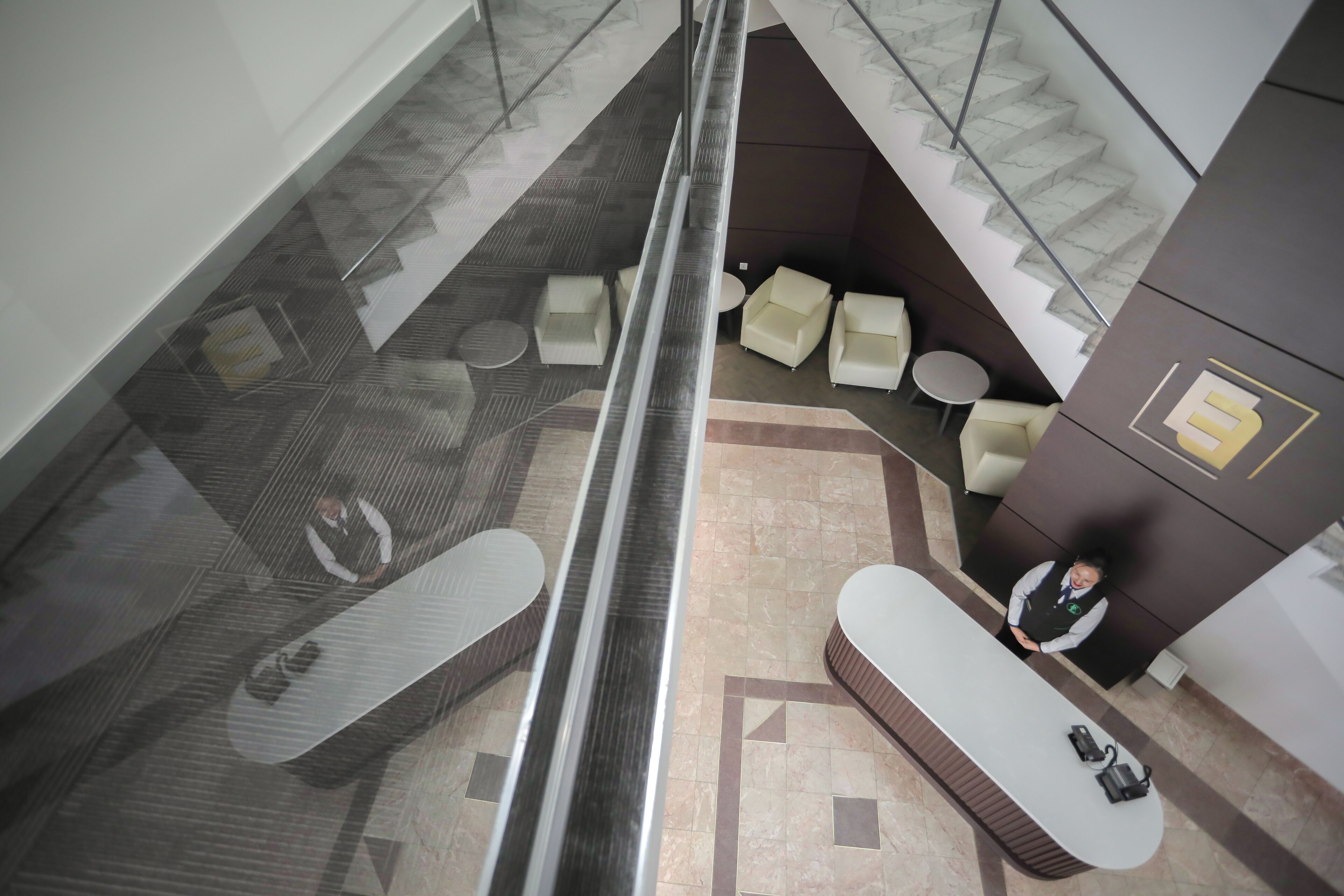 A modern hotel lobby with a glass elevator, seating area with sofas, and a reception desk.