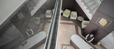 A modern hotel lobby with a glass elevator, seating area with sofas, and a reception desk.
