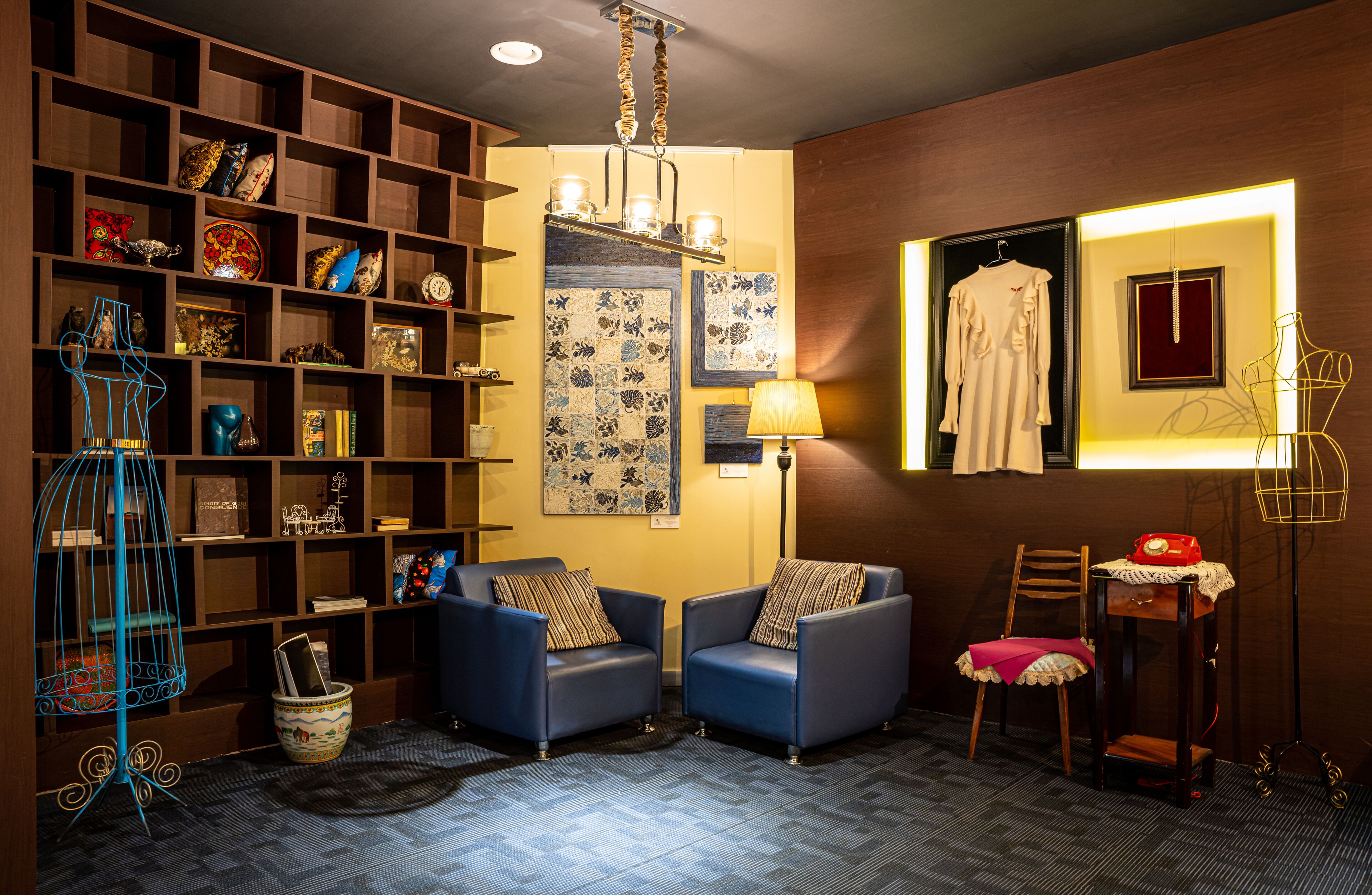 Una habitación acogedora con una estantería de madera, dos sillones azules, una lámpara de pie y una mesita con un teléfono rojo.