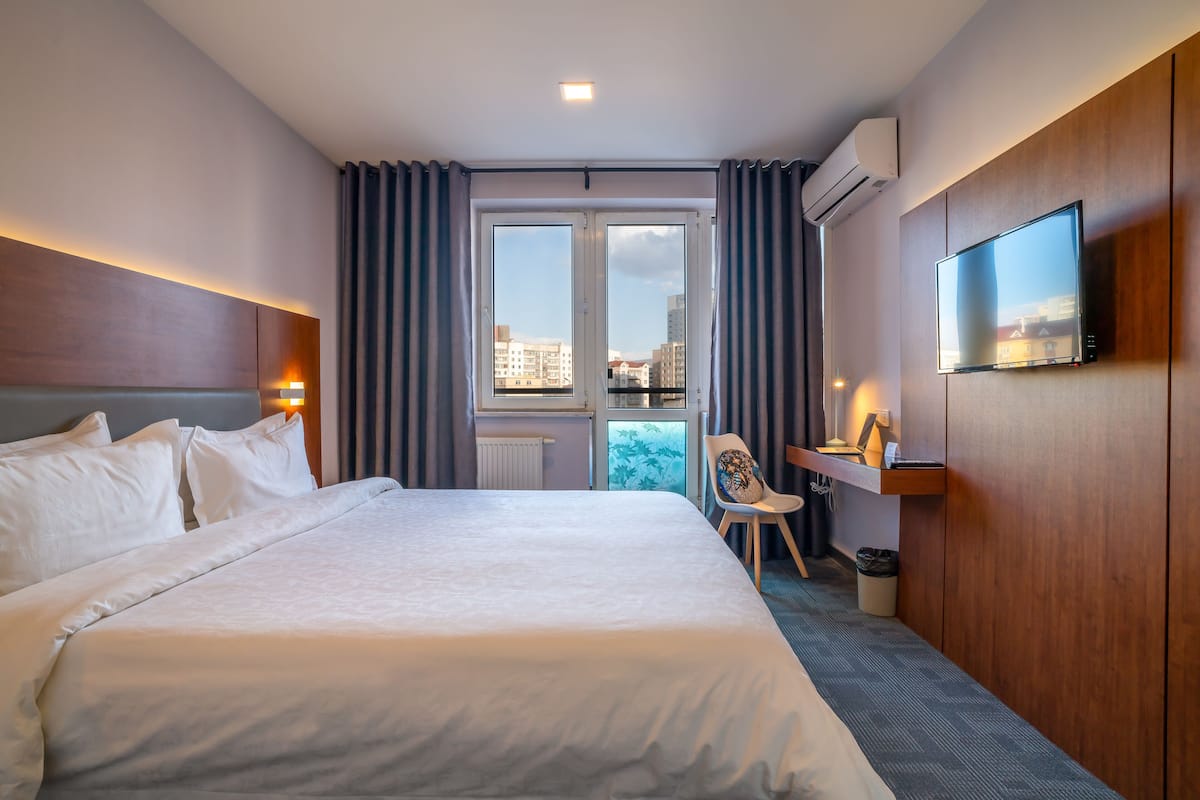 A hotel room with a large bed, a desk, a chair, a television, and a view of the city through the window.