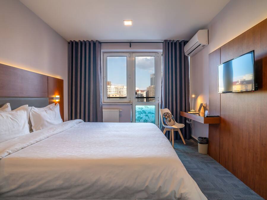 A hotel room with a large bed, a desk, a chair, a television, and a view of the city through the window.