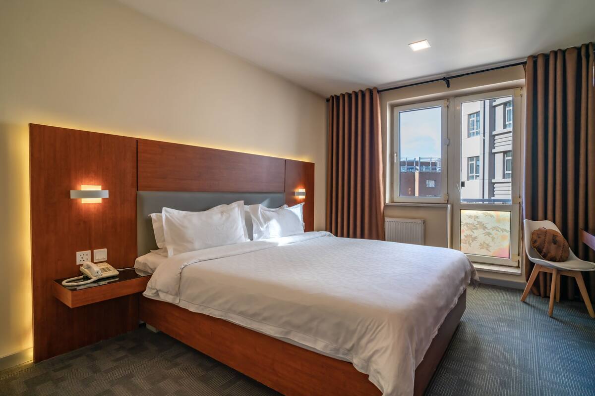 A hotel room with a large bed, a bedside telephone, and a window with curtains.