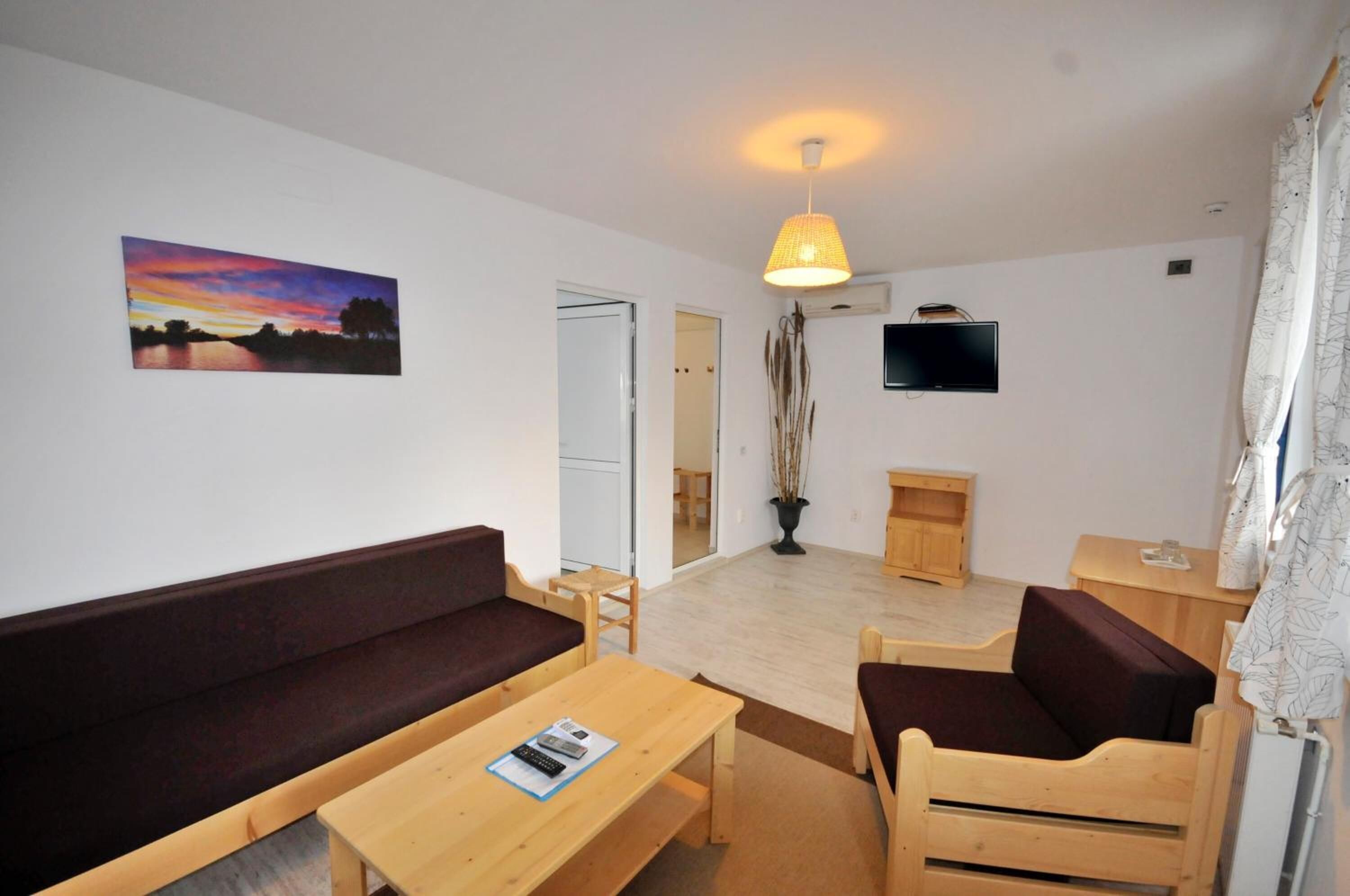 A cozy living room with a sofa, wooden chairs, a table, a TV, and a painting of a sunset.