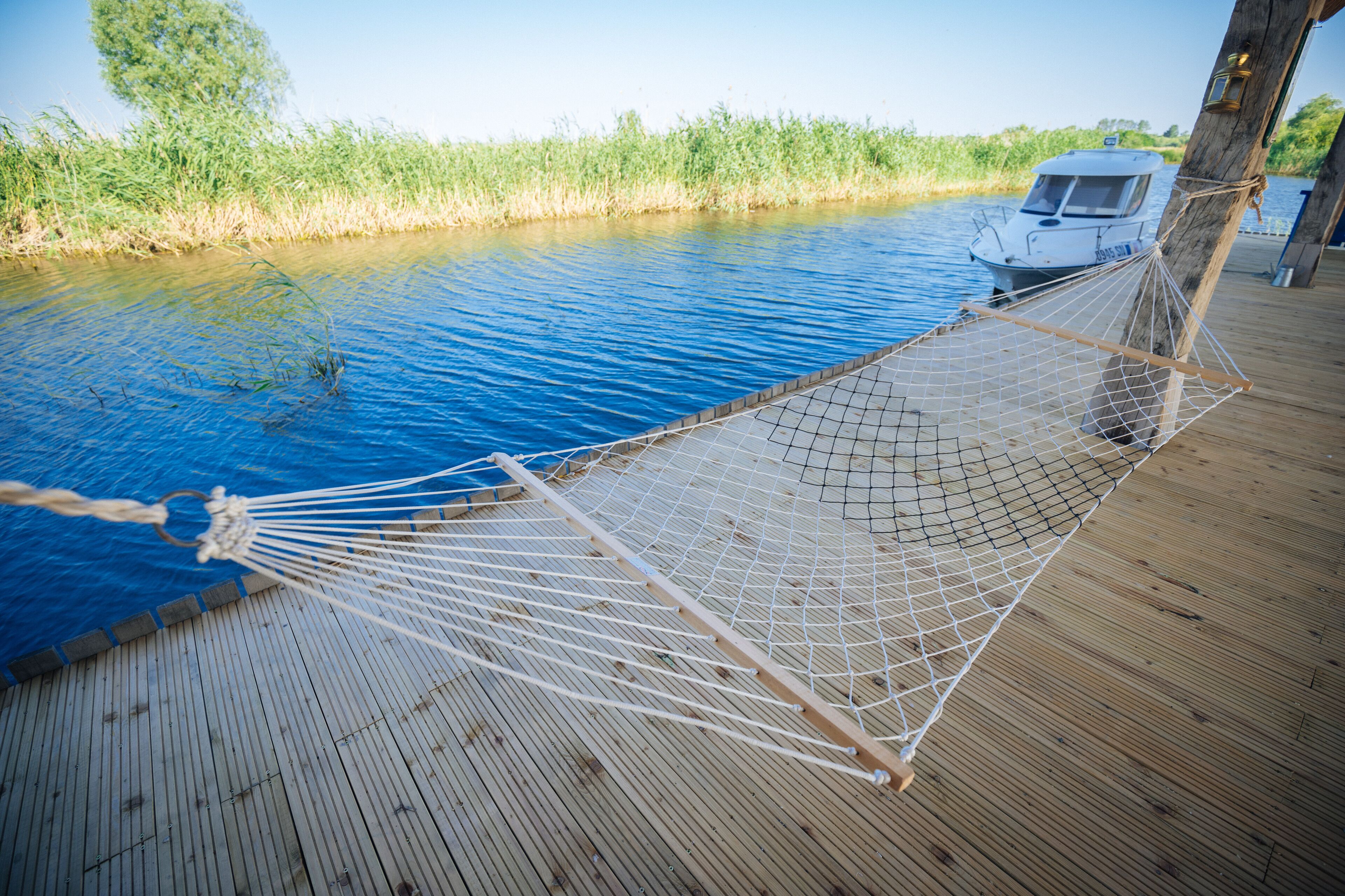 Un hamac sur une terrasse en bois, au bord d’une rivière paisible, avec un bateau en arrière-plan.