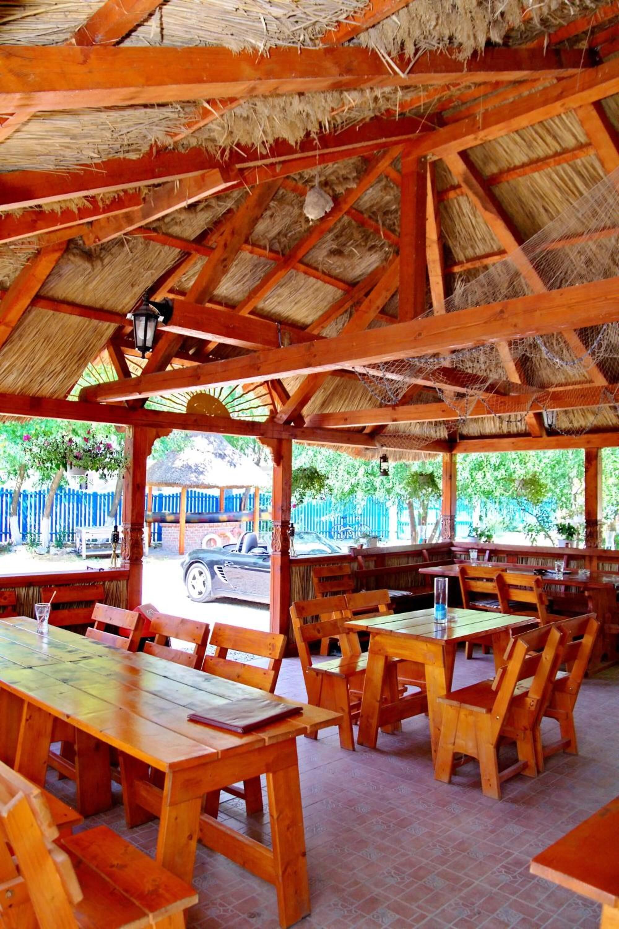 An outdoor wooden dining area with tables and benches, a thatched roof, and a view of a street with a car and a fence.