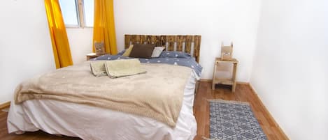 Un dormitorio con cabecera de madera, cama con manta color beige, mesita de noche y alfombra estampada.