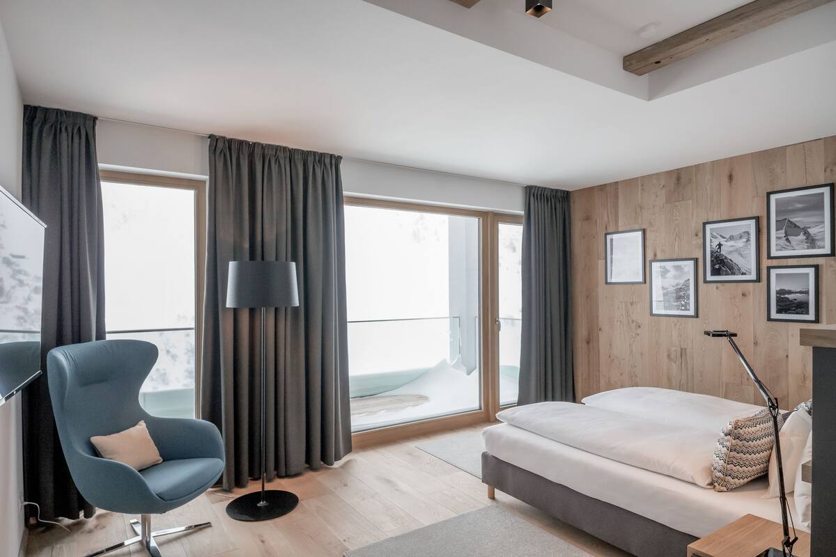 Een moderne hotelkamer met een groot bed, een blauwe fauteuil, een staande lamp en uitzicht op het omliggende landschap door grote ramen.