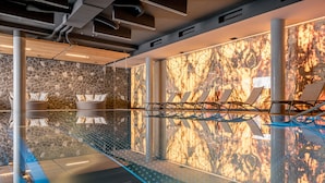 Een modern binnenzwembad met een spiegelende vloer, een stenen muur en ligstoelen.