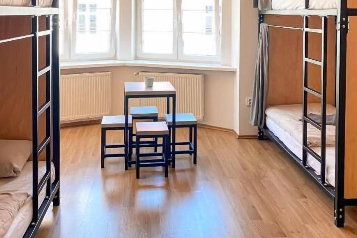 A dormitory room with bunk beds, a small table, and chairs. There are large windows providing natural light.