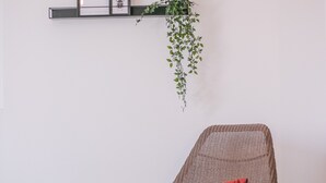 Une pièce avec un fauteuil en osier doté d’un coussin rouge, un lit recouvert d’un drap blanc, un plancher en bois, une étagère murale ornée de tableaux encadrés et de plantes, et une plante suspendue.